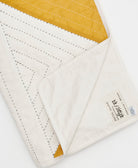 Close-up of a quilt with white and mustard yellow fabric, featuring Anchal's artisan signed tag