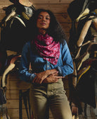 Woman wearing a pink silk square scarf and blue denim shirt standing among horse saddles in a barn setting