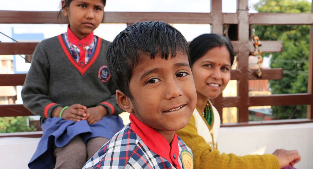 Two Indian children dressed in school uniform with an Indian woman, smiling at the camera