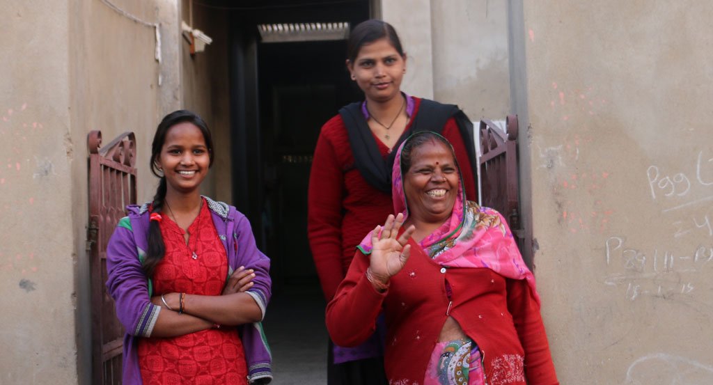 three Indian women outside of their home waving at the camera