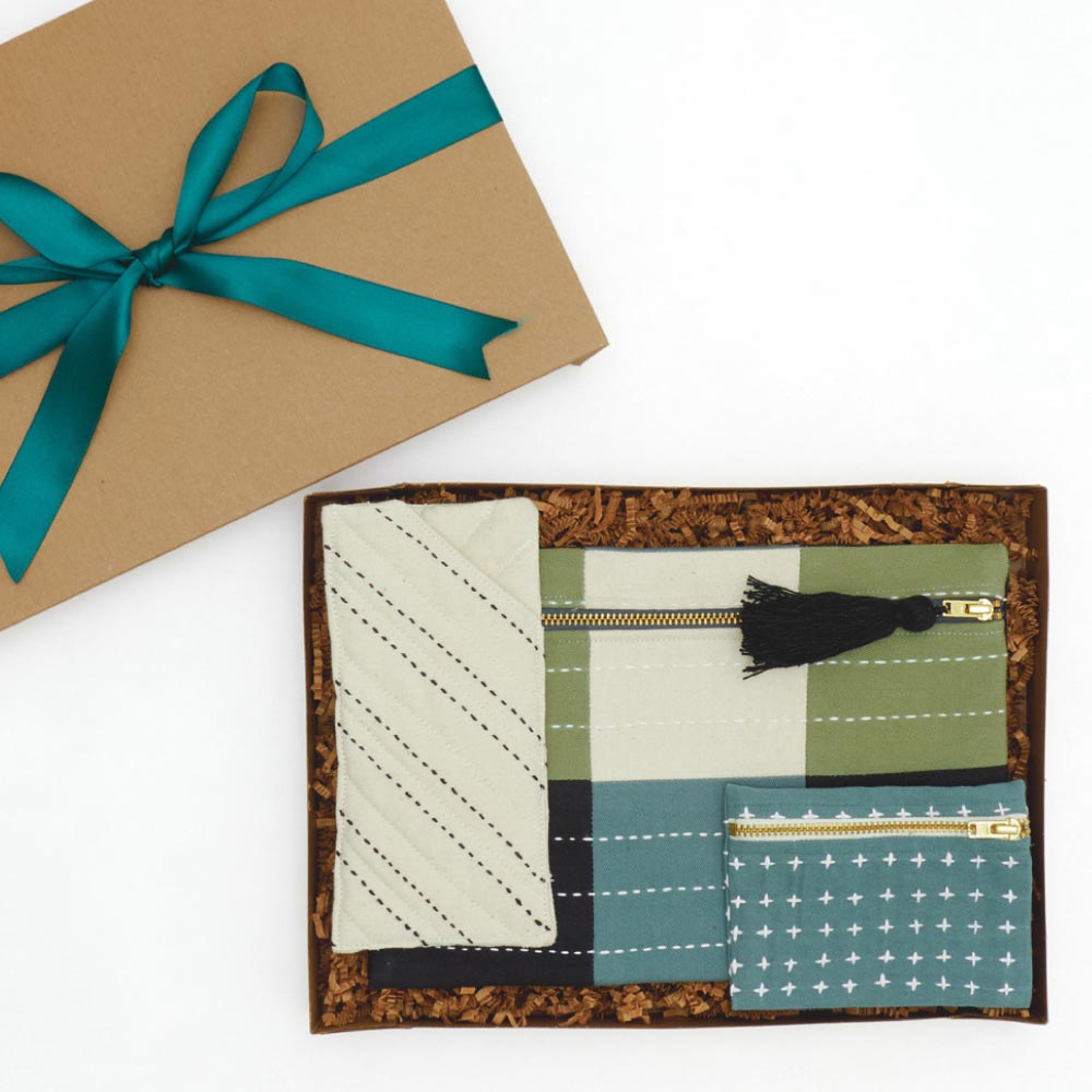 brown gift box with a green ribbon containing a cool-toned checkered pouch, coin purse and glasses case
