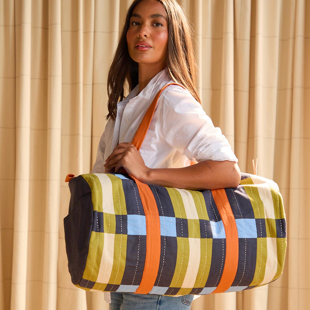 woman carrying a navy blue, orange and green patterned duffle bag over her shoulder wearing a white button up top