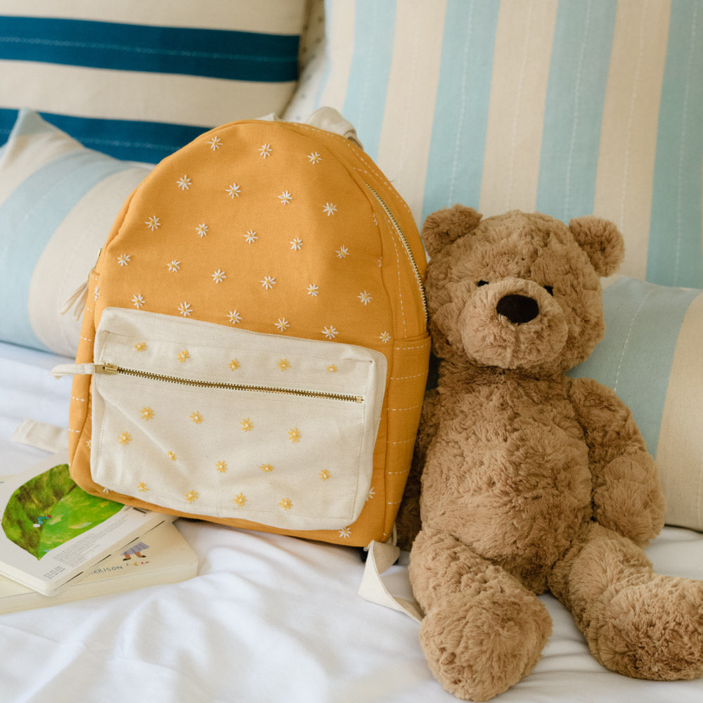 mustard yellow mini backpack with daisy embroidery sitting on blue and white bed with brown teddy bear
