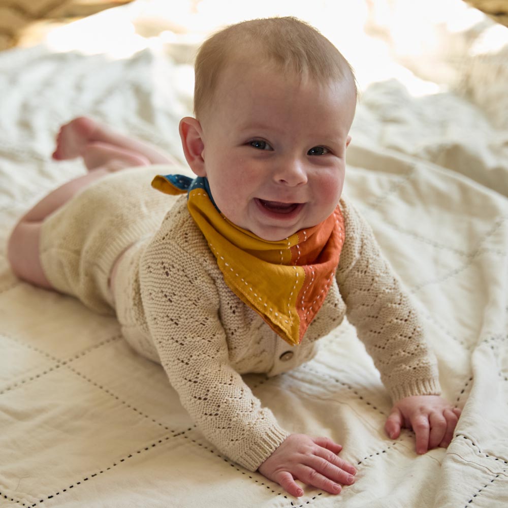 baby girl wearing ivory sweater and colorblock reversible drool bib in shades of blue, yellow, orange & pink