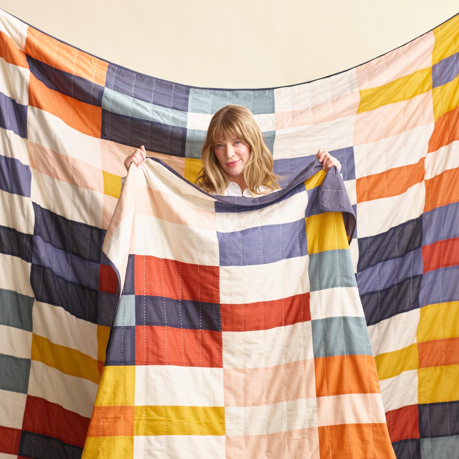 woman holding up colorful checkered quilt with patchwork quilt in background