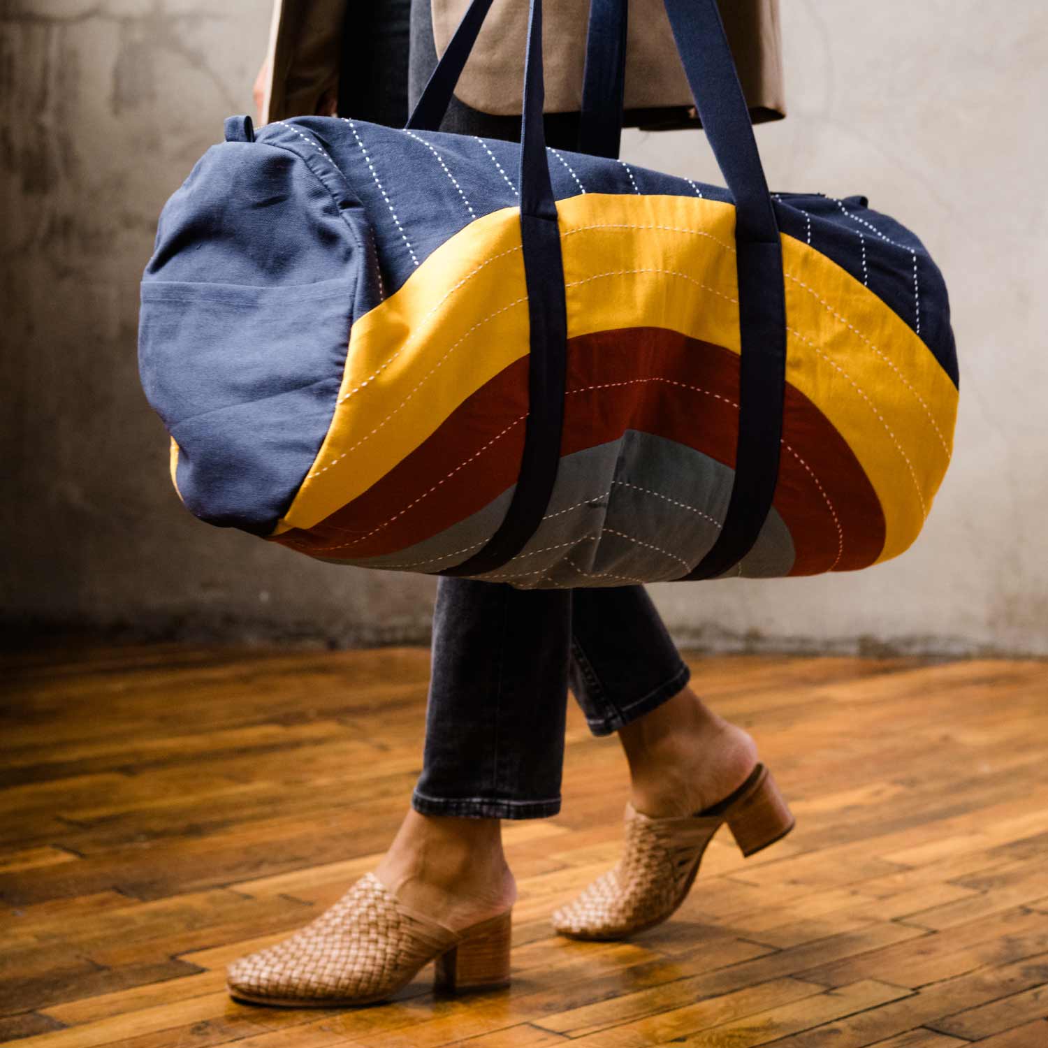 woman holding barrel shaped canvas duffle bag in a rainbow design with navy blue straps