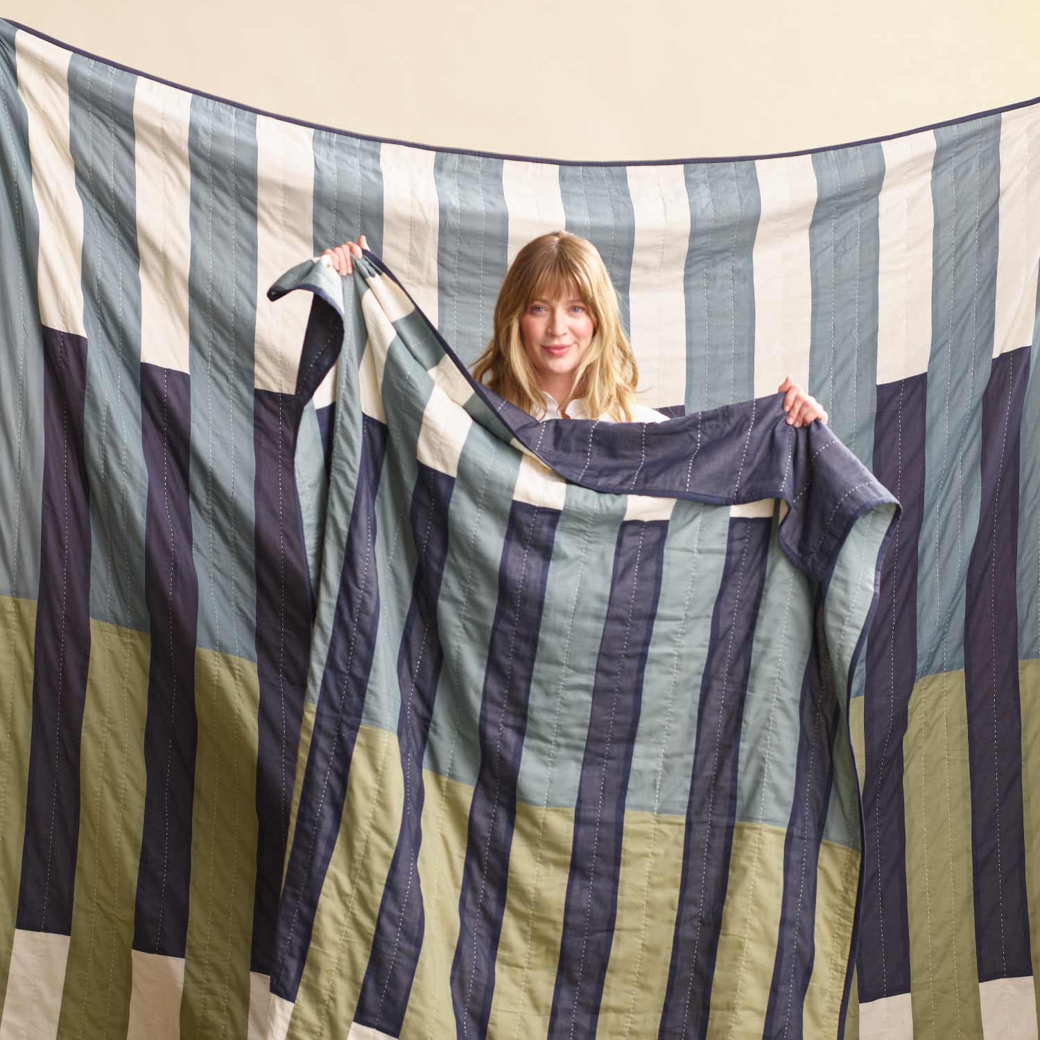 woman holding striped patchwork quilt in front of larger striped bedding quilt in shades of blue and green