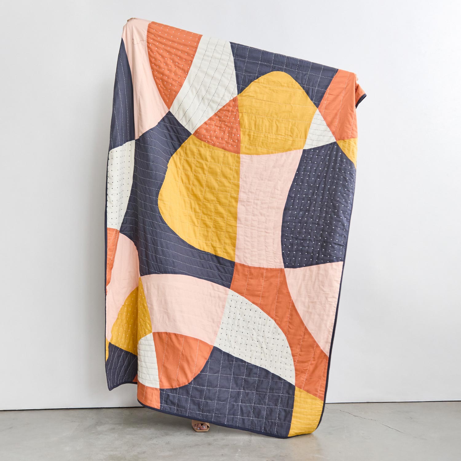 abstract patchwork quilt in navy blue, yellow, orange and pink being held up against an all white wall