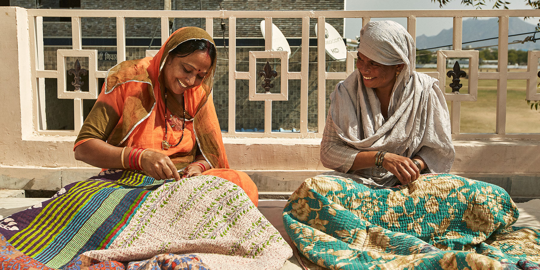 two women artisans in Ajmer, India stitching colorful kantha quilts on balcony and smiling