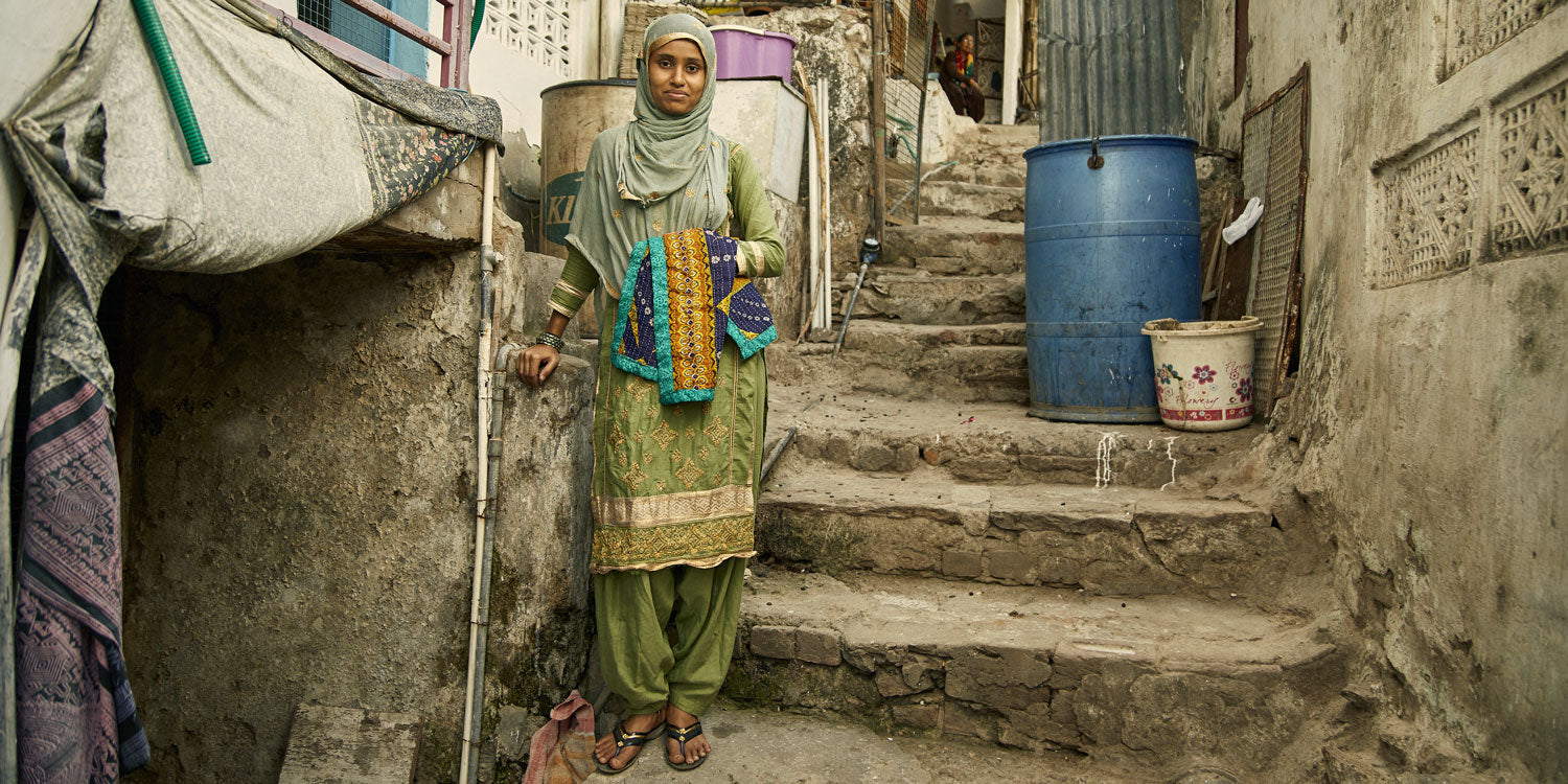 Anchal artisan standing on stone stairs in front of her home holding an Anchal kantha quilt she made