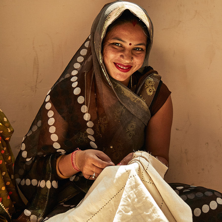 Anchal artisan smiling wearing brown sari and hand-stitching a patchwork quilt