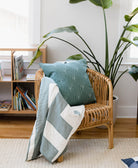 embroidered spruce green throw pillow in modern rattan chair next to modern bookshelf