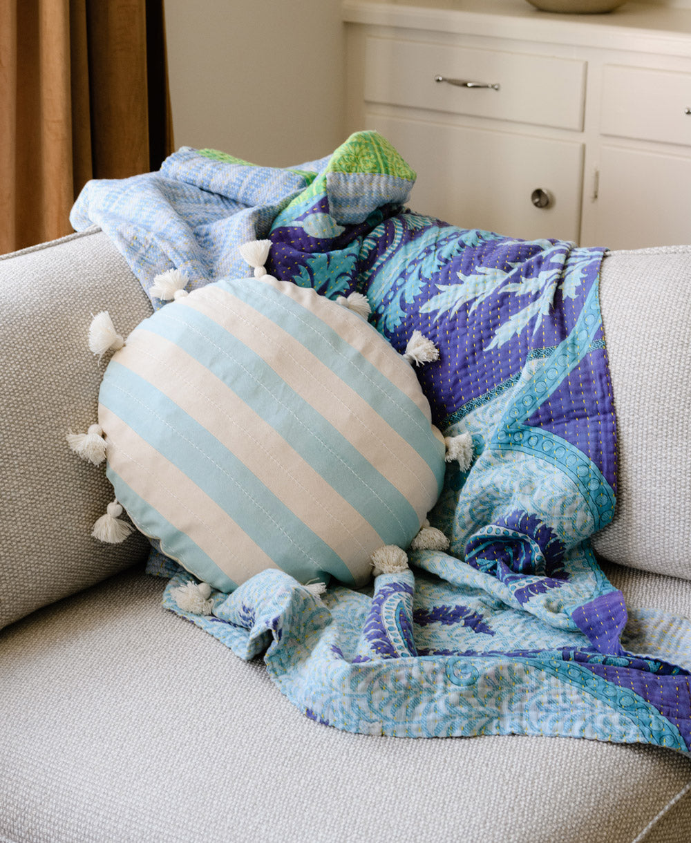 light blue striped round pillow with white tassels on modern grey sofa with coordinating blue & purple kantha quilt