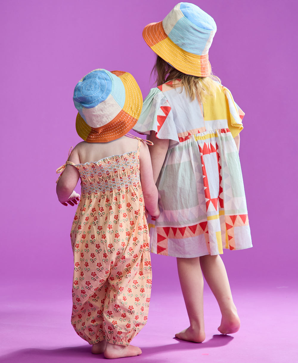 two little girls holding hands wearing checkered bucket hats while holding hands on pink backdrop