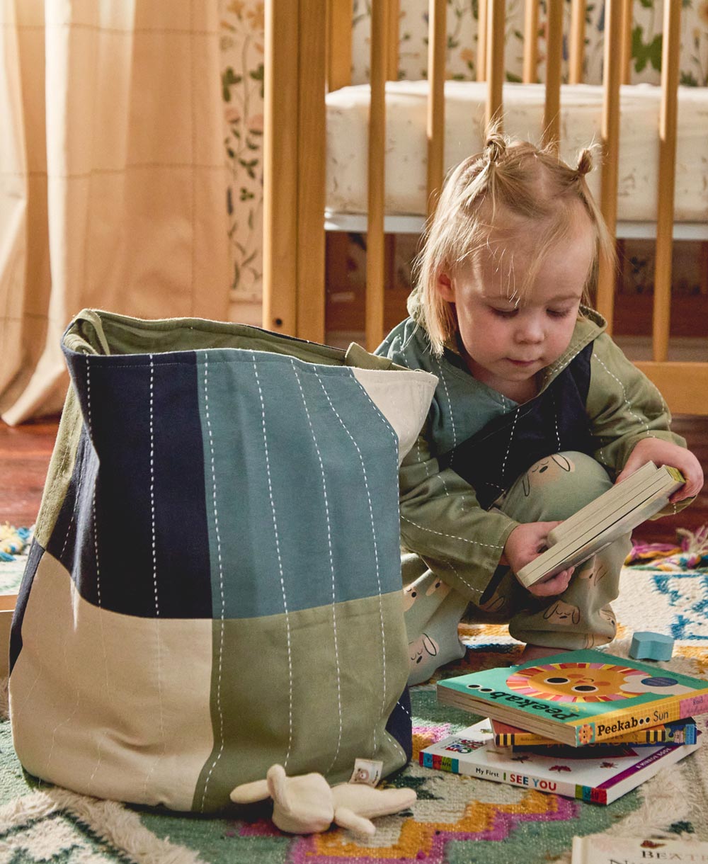 toddler wearing colorblock hoodie putting books into round fabric storage bin in checkered design in bedroom