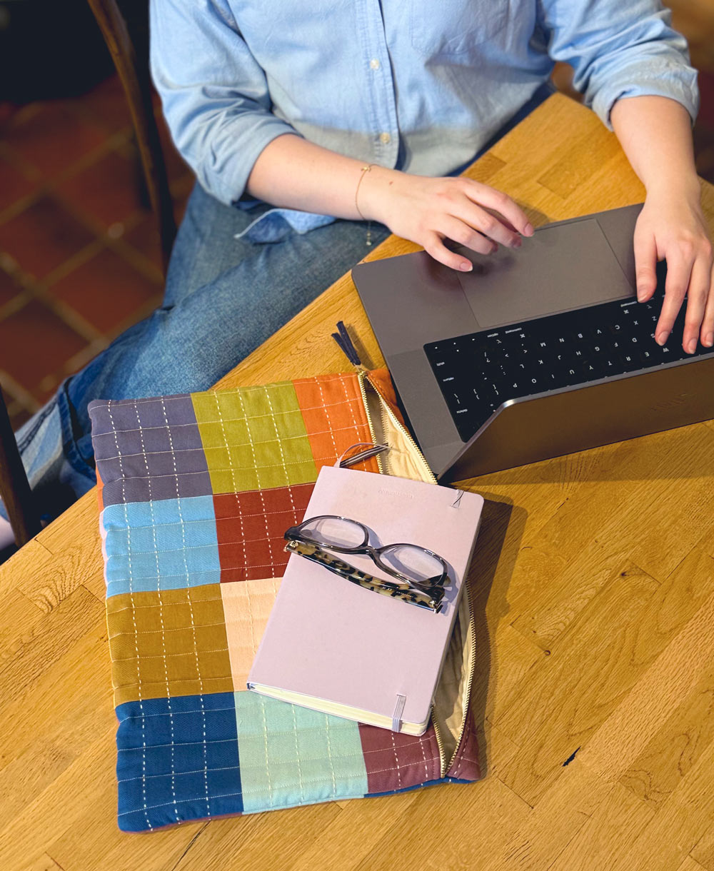 Person using a laptop on a wooden table with a colorful quilted mat and notebook.