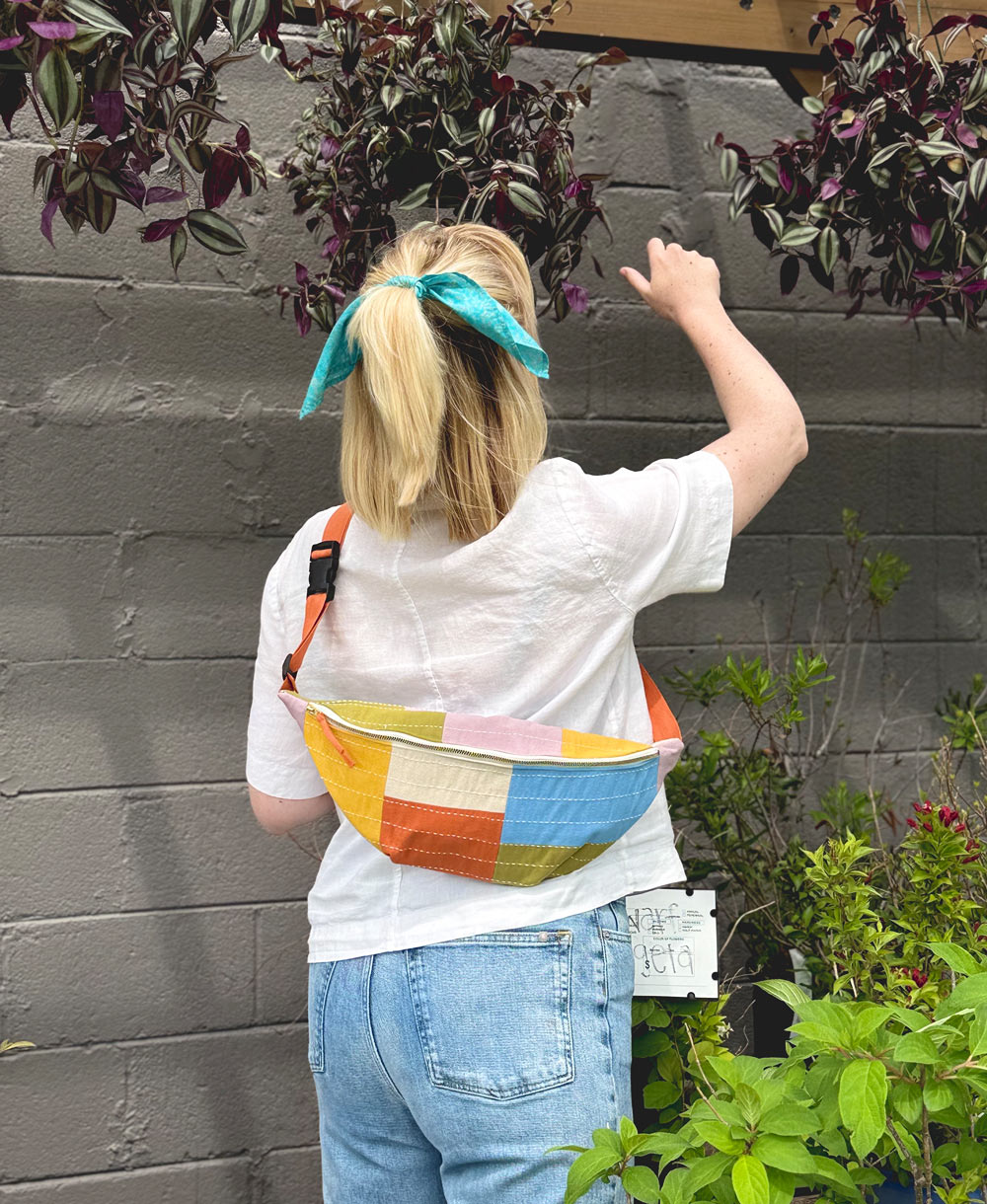 woman wearing checkered sling bag in cloud with a white button up shirt and jeans reaching for a purple potted plant