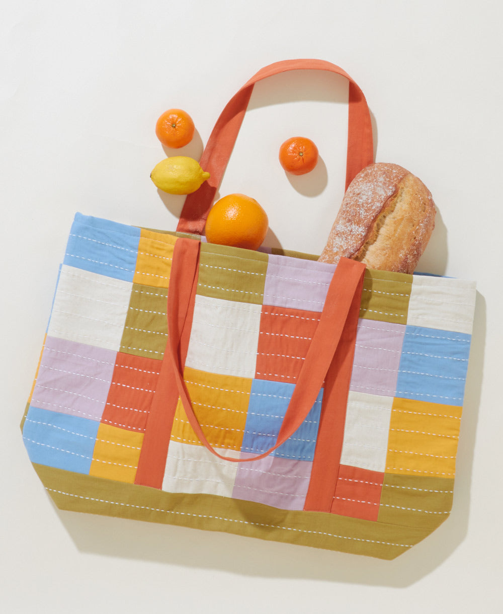 colorful checkered canvas tote bag with orange straps with bread and fruit spilling out for the farmer's market