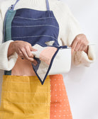 woman wearing colorblock kitchen apron holding a white pot with a matching quilted pot holder in navy and pink