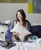 woman sitting on all white bed holding white small pillow with black cross-stitching pattern