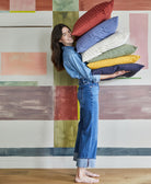 woman in all denim outfit holding a stack of solid color square pillows in a rainbow of hues against pastel wall mural