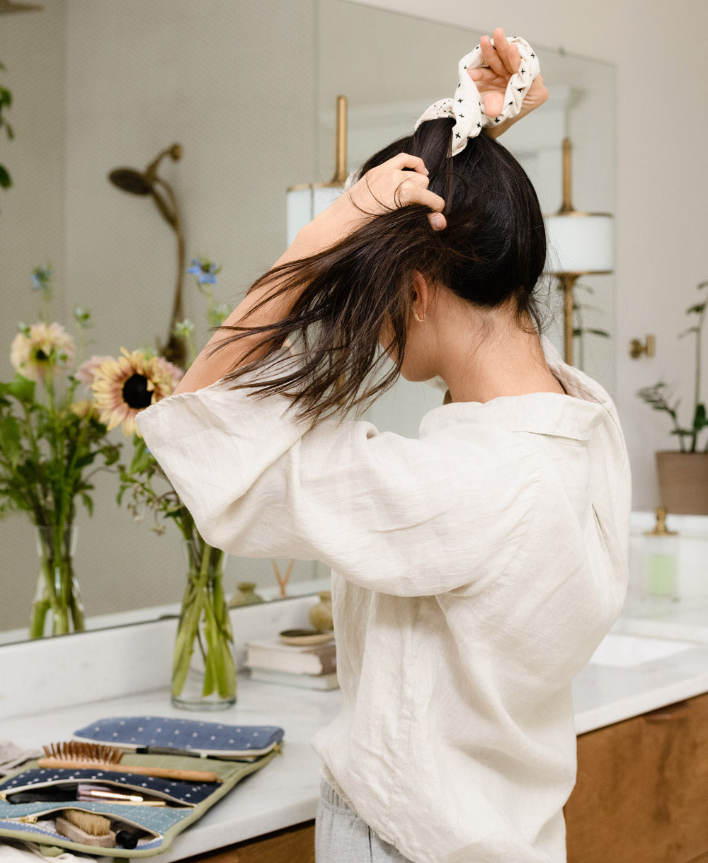 woman in matching loungewear linen set tying her hair in a ponytail with an ivory embroidered scrunchie