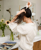 woman in matching loungewear linen set tying her hair in a ponytail with an ivory embroidered scrunchie