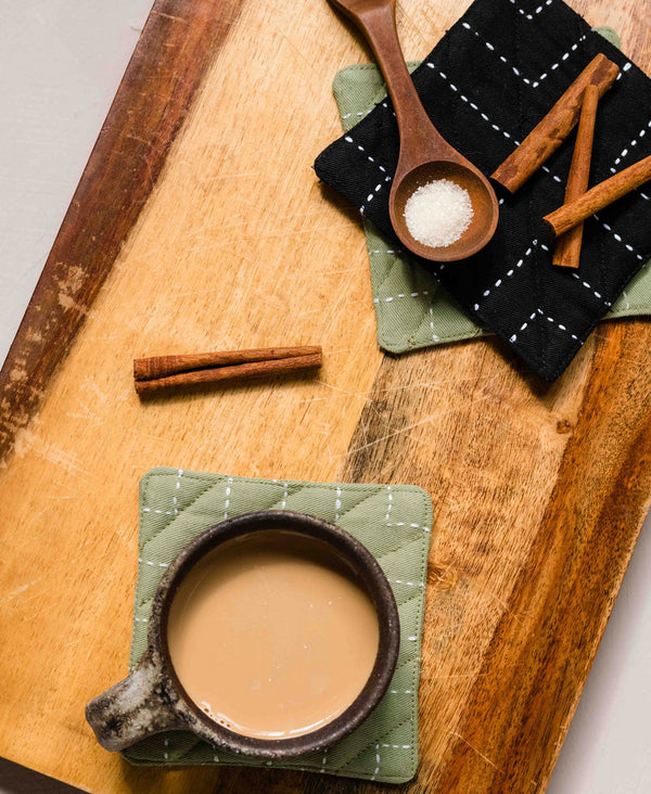 black and sage green hand-embroidered coasters on wooden cutting board with cup of chai tea