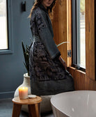 cream glass jar candle sitting on wooden bathroom stool with woman in grey silk robe standing behind