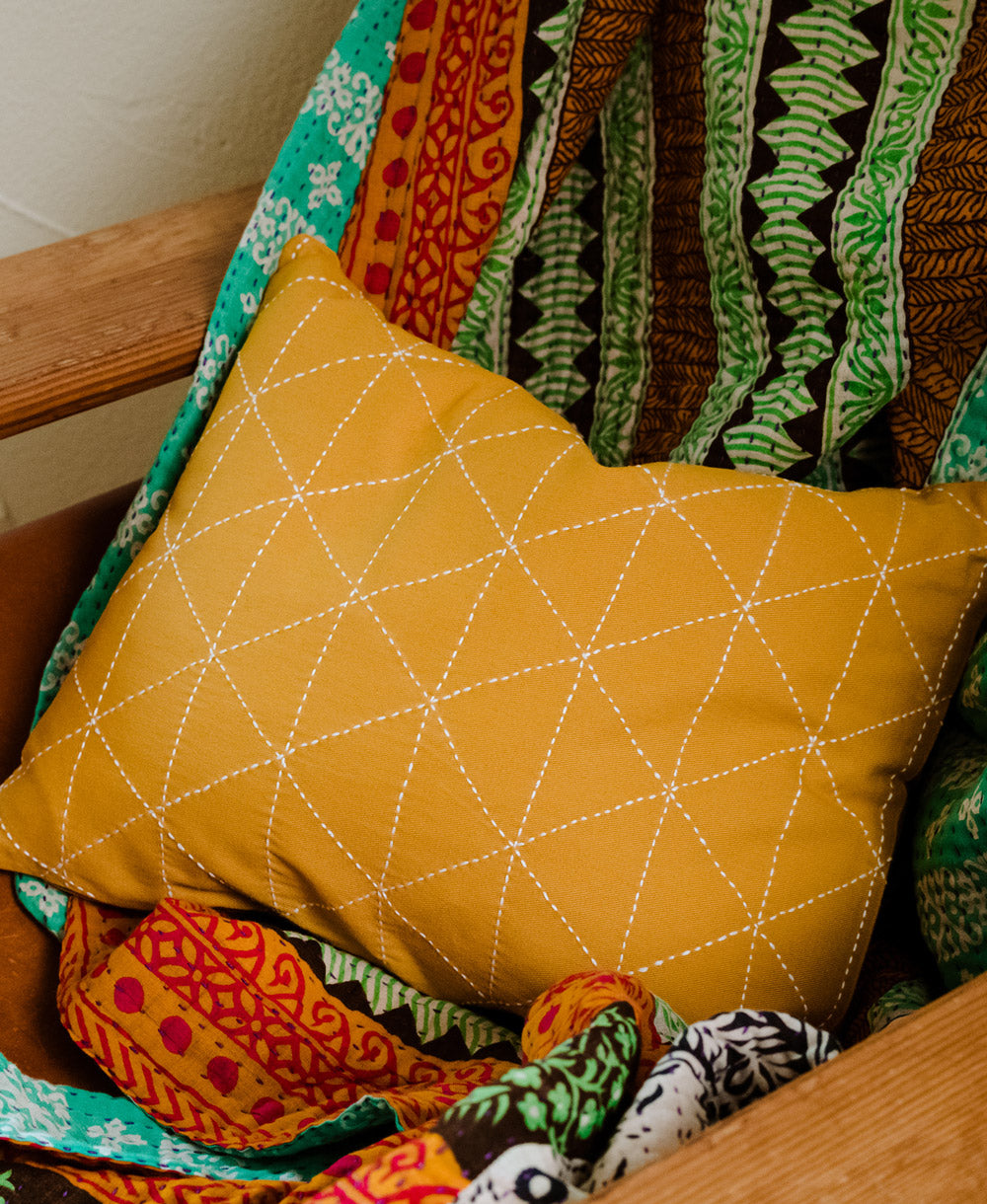 small throw pillow in mustard yellow with white stitching in chair with mismatched vintage kantha quilt