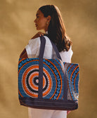 woman holding a colorful canvas tote bag over her shoulder wearing an all white linen outfit against a beige background