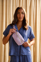 woman holding lilac crossbody belt bag in front of beige backdrop