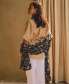 woman wearing a black and grey floral long cotton scarf against a beige background with ivory grid-stitch curtains