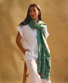woman wearing a long green cotton scarf sitting on a wooden stool against a beige background
