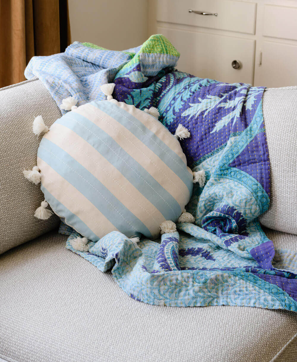 blue and purple kantha quilt draped over grey sofa with round striped blue tassel pillow