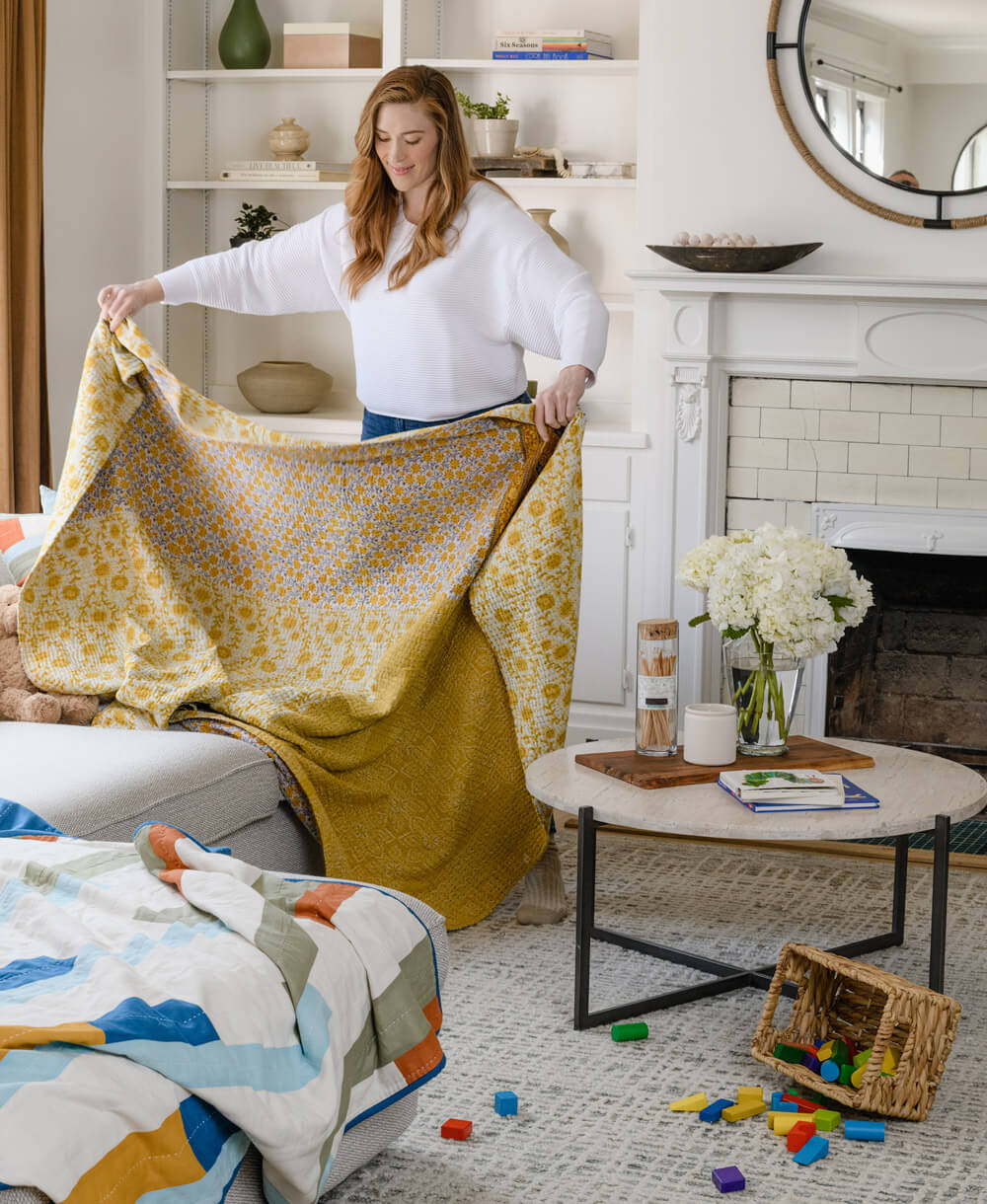 woman folding yellow floral kantha quilt in living room onto modern grey sofa