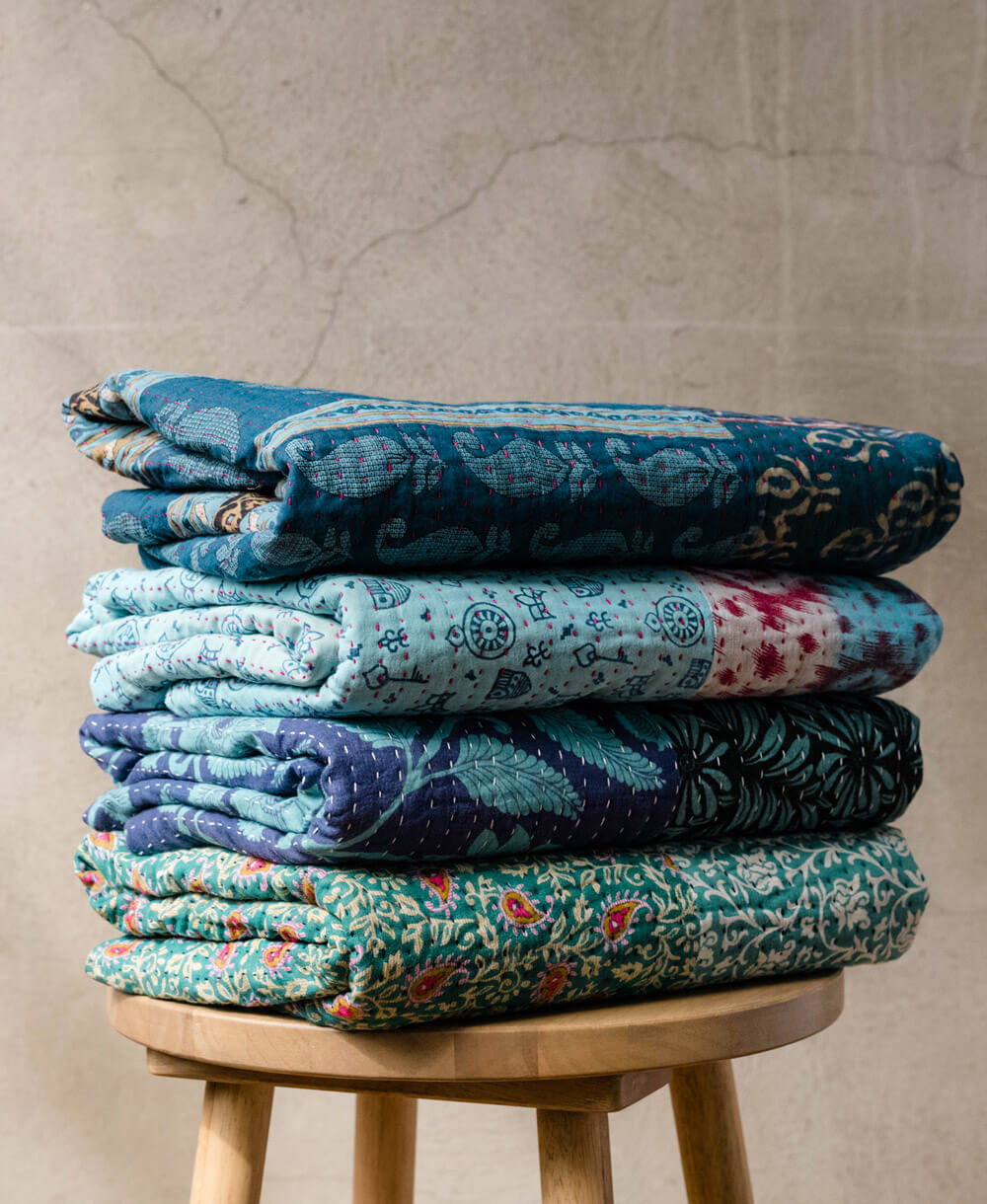 stack of blue kantha quilts handmade in India by women artisans on natural wooded stool