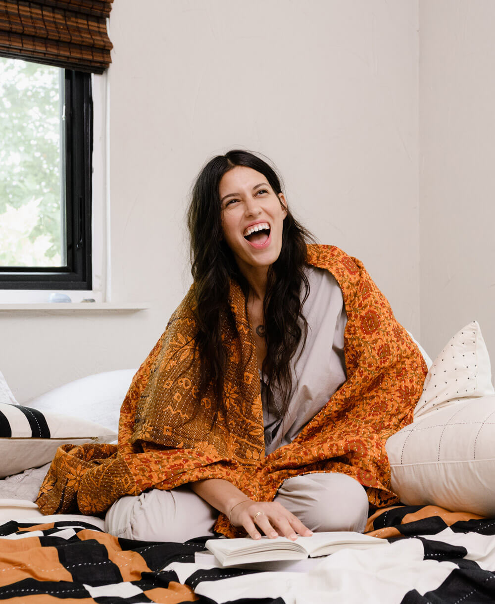 woman with long dark hair draped in a rust orange kantha quilt sitting on bed