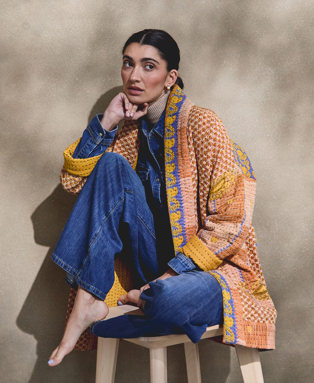 woman sitting on wooden stool in all denim outfit accented with orange & blue quilted jacket made from vintage saris