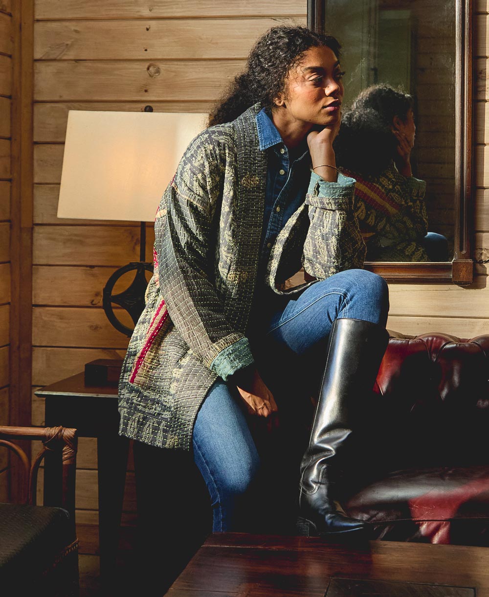 Woman sitting on a leather couch in a cozy room with wooden walls, wearing an open front quilted jacket in dark grey