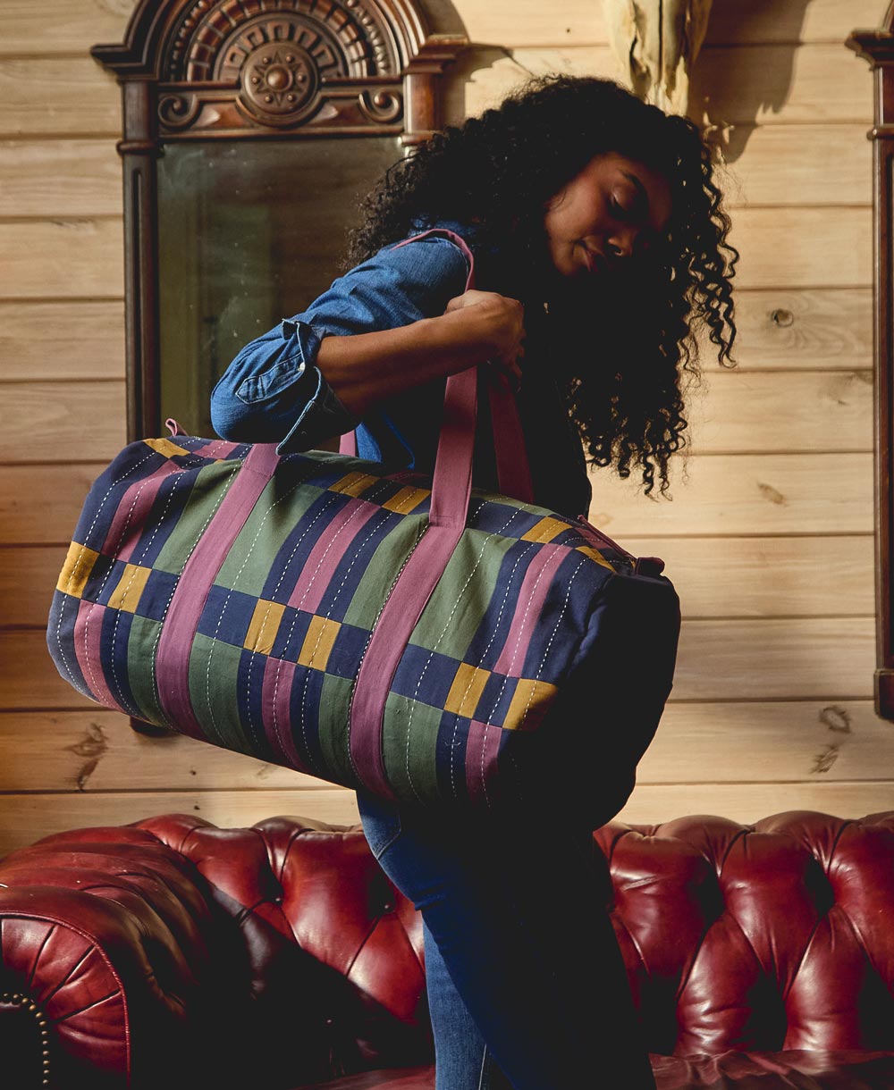 Woman holding a colorful striped duffel bag in a room with wooden walls and a red leather couch