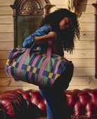 Woman holding a colorful striped duffel bag in a room with wooden walls and a red leather couch