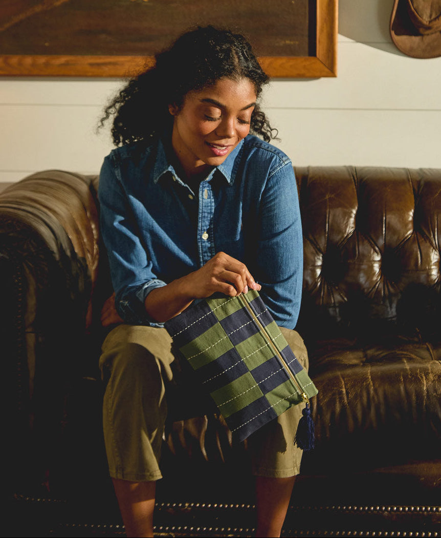 Woman sitting on a brown leather couch holding a plaid bag, with a painting of a bird and a cowboy hat in the background