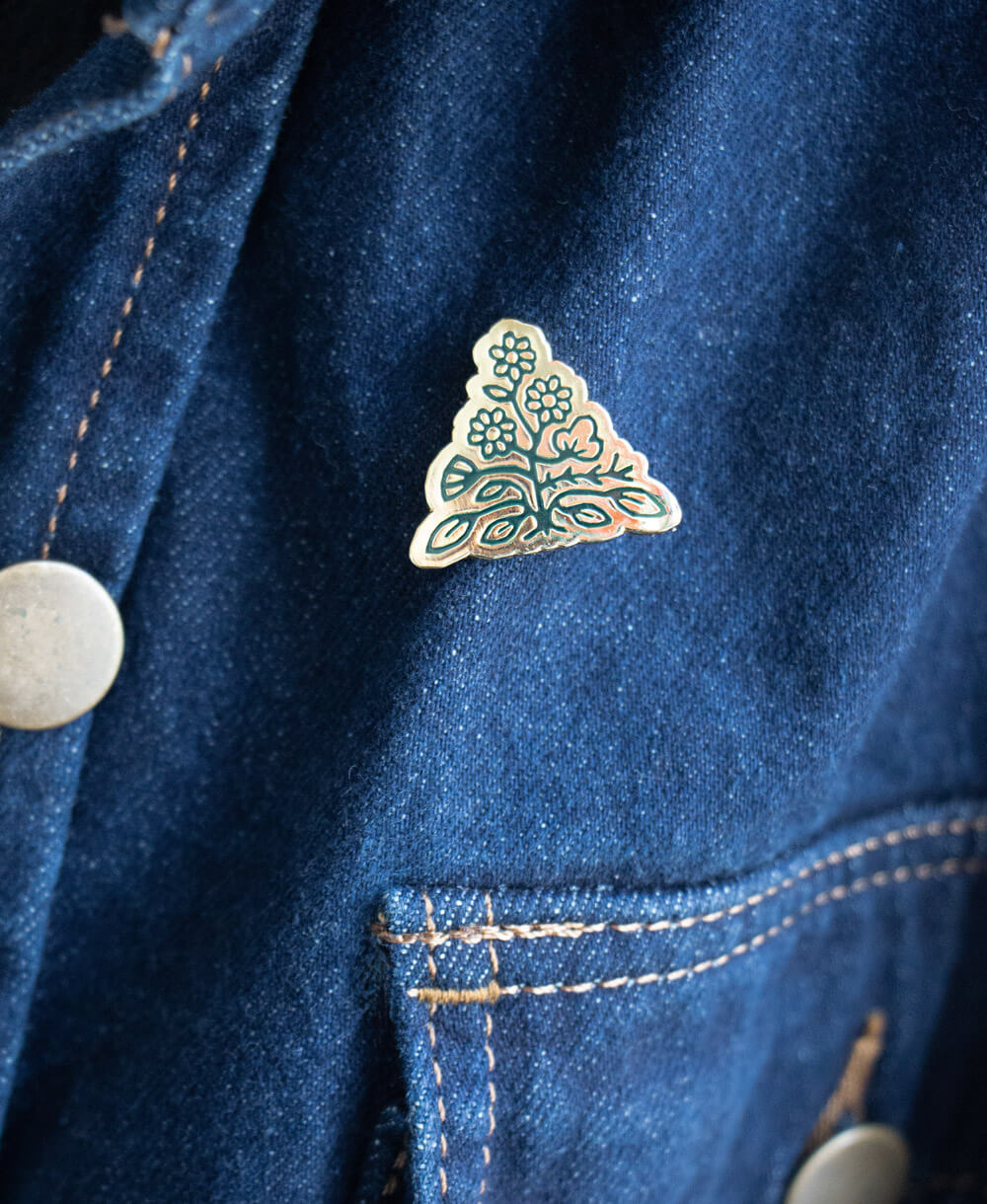 gold hard enamel pin in a triangle shape featuring Anchal's brand mark pinned on dark denim jacket