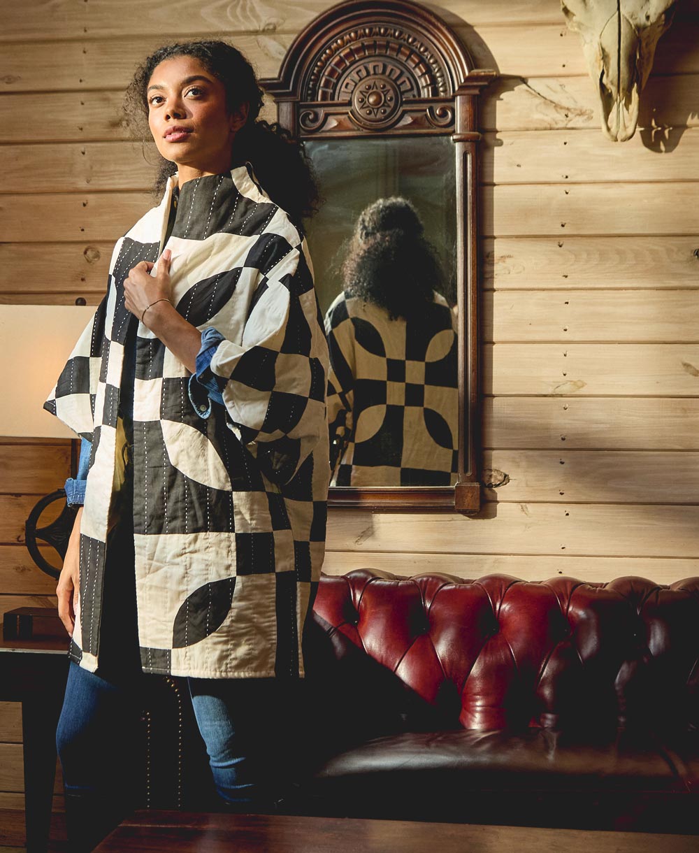 Woman wearing a black and white patterned quilted jacket standing in a room with wooden walls and a red leather couch.