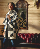 Woman wearing a black and white patterned quilted jacket standing in a room with wooden walls and a red leather couch.