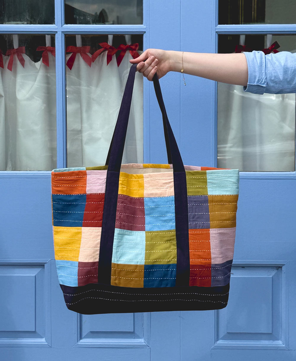 Colorful checkered tote bag held by a hand in front of a blue door with white curtains.