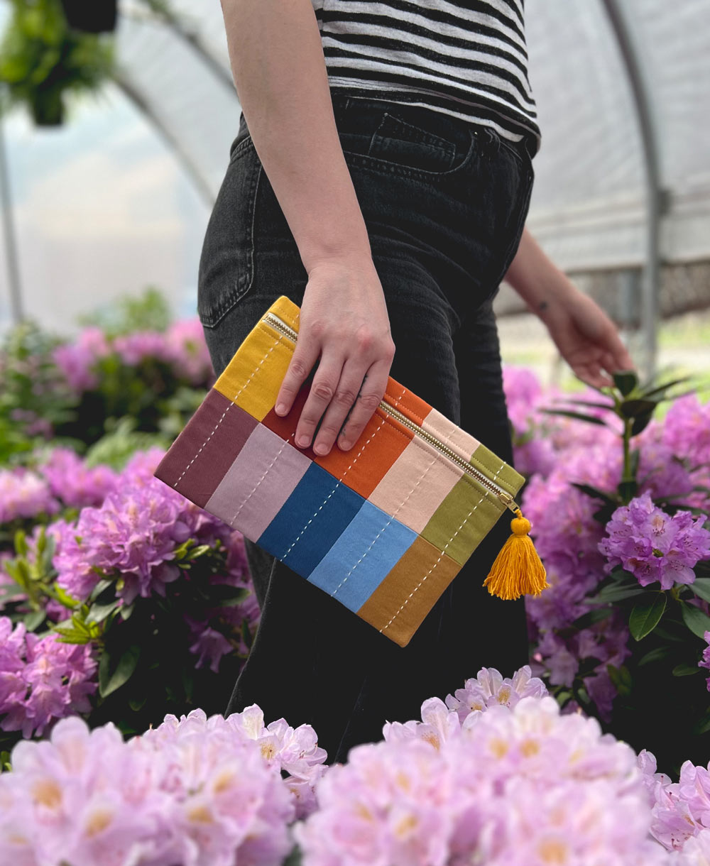 person in black jeans and a striped black and white shirt holding a colorful checkered pouch with a yellow tassel zipper pull