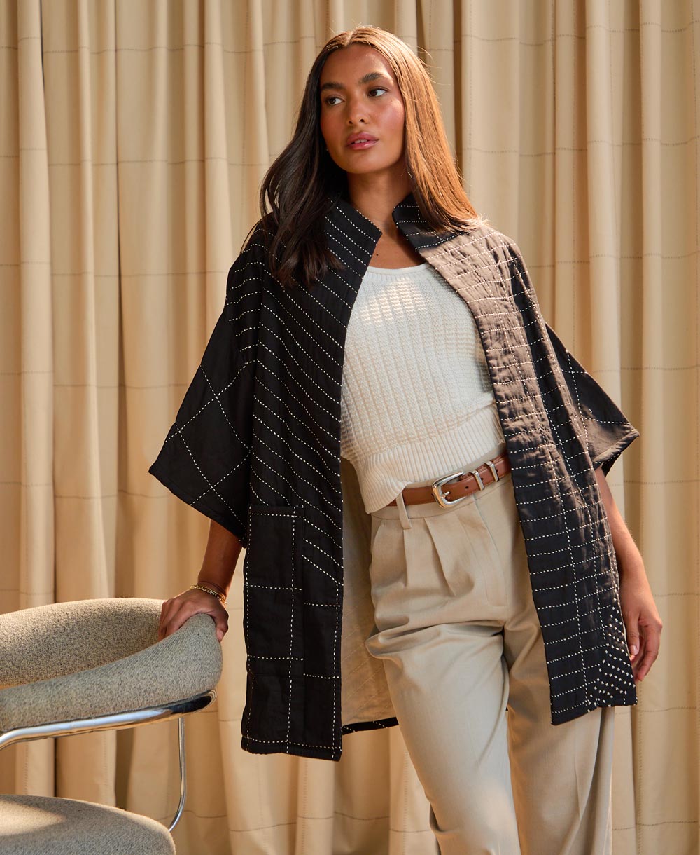 woman leaning on chair wearing black quilted cocoon jacket in front of beige backdrop