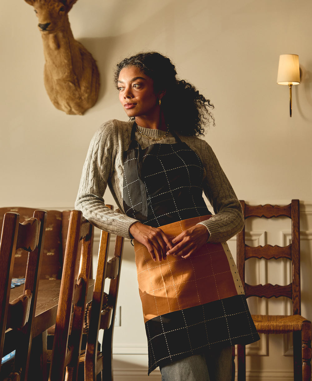 Woman wearing a checkered apron standing in a room with wooden chairs and a deer head on the wall